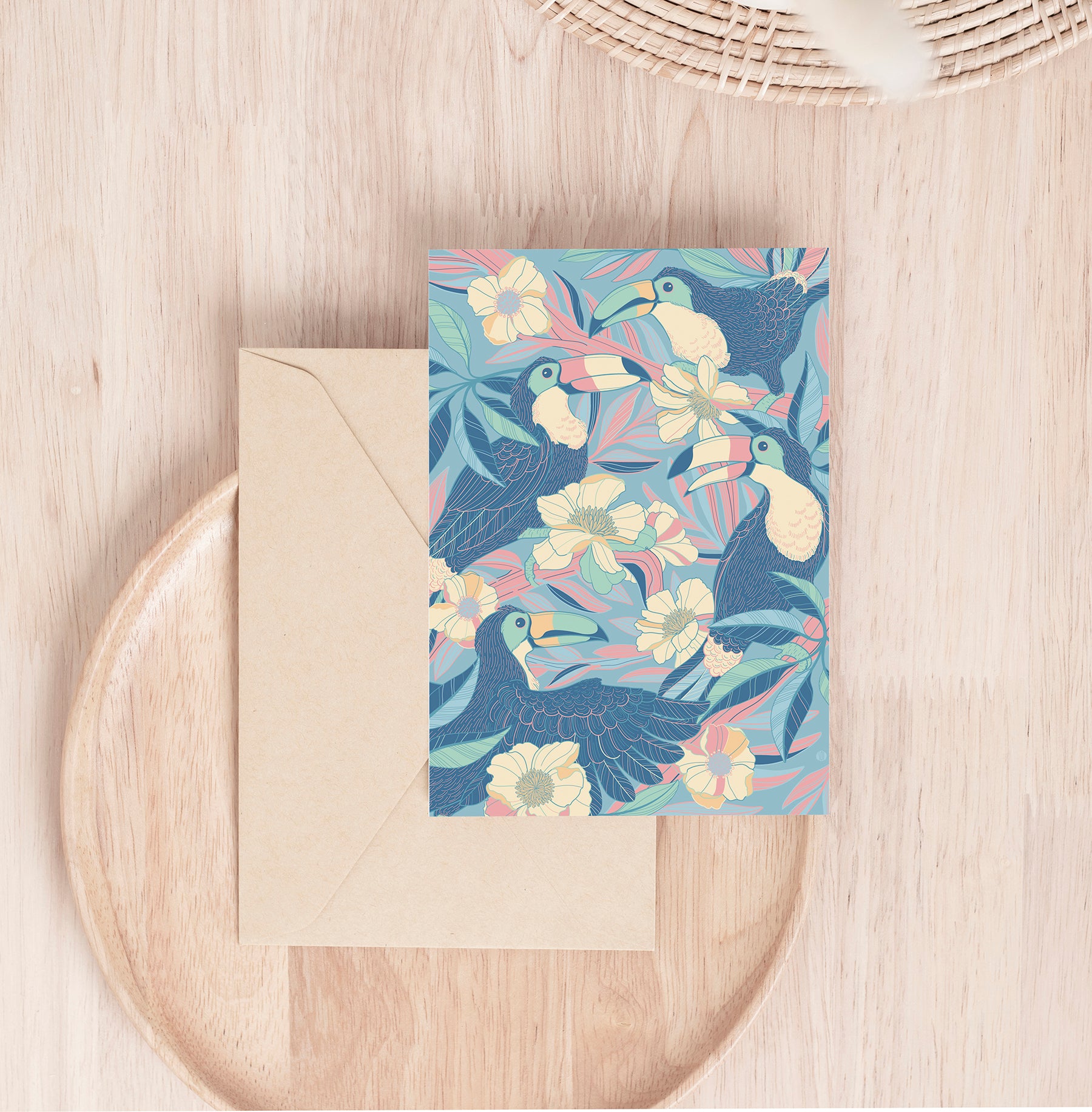 Colorful toucan and floral illustration on a card, set on a wooden surface with a beige envelope.