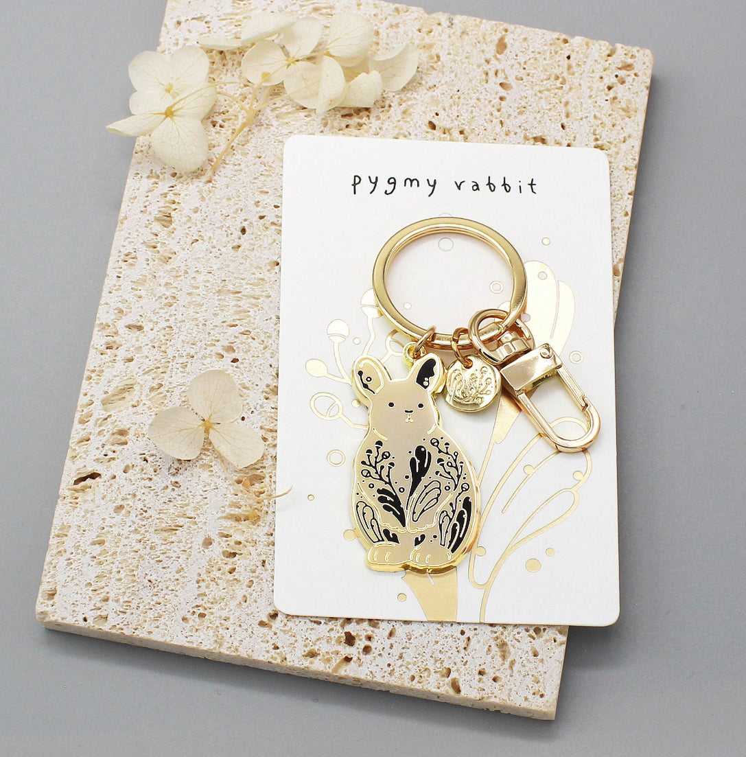 Pygmy rabbit keychain with habitat design, displayed on decorative card surrounded by flowers.