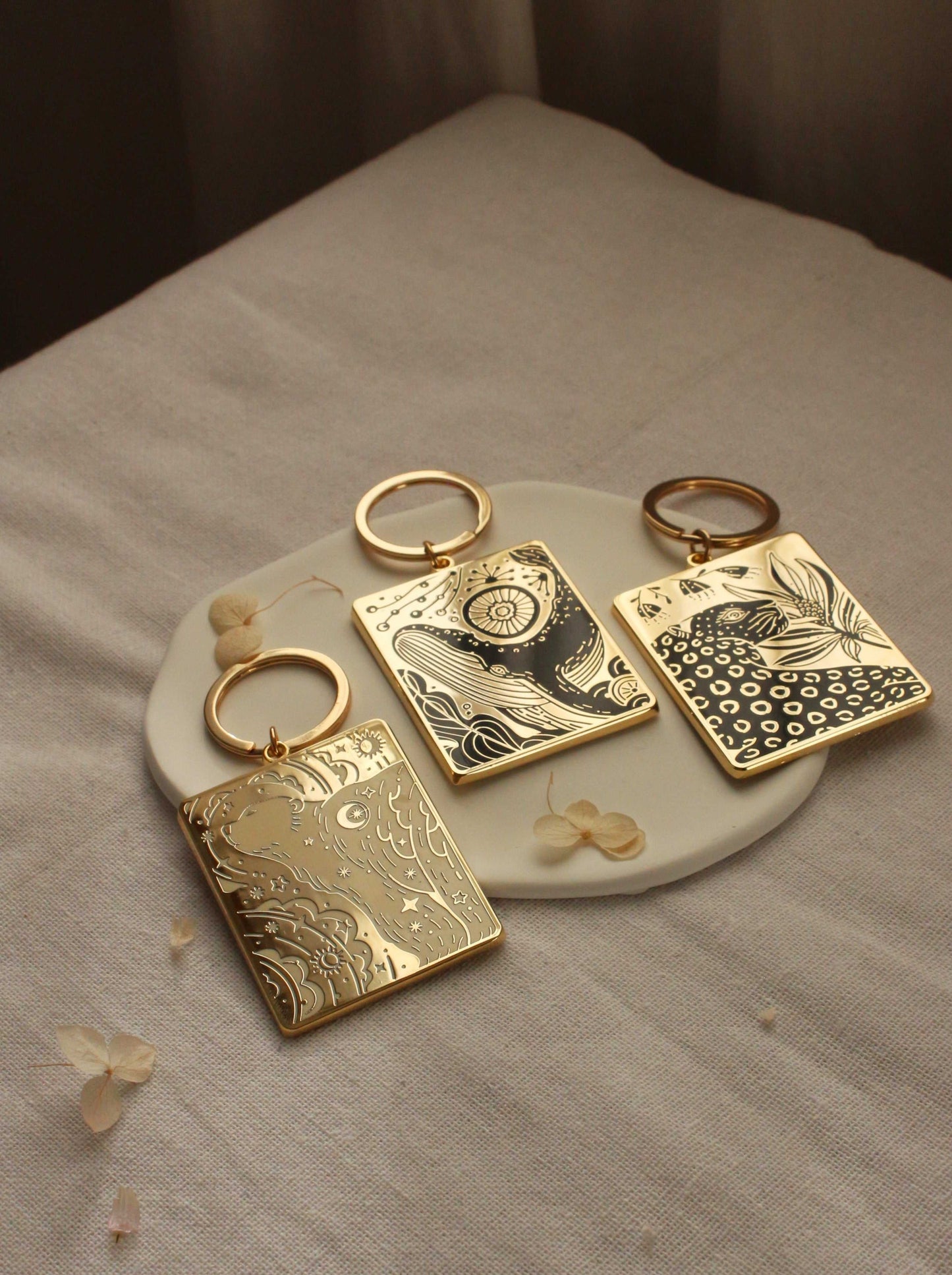 Set of three gold-plated enamel keychains with intricate designs displayed on a ceramic plate.