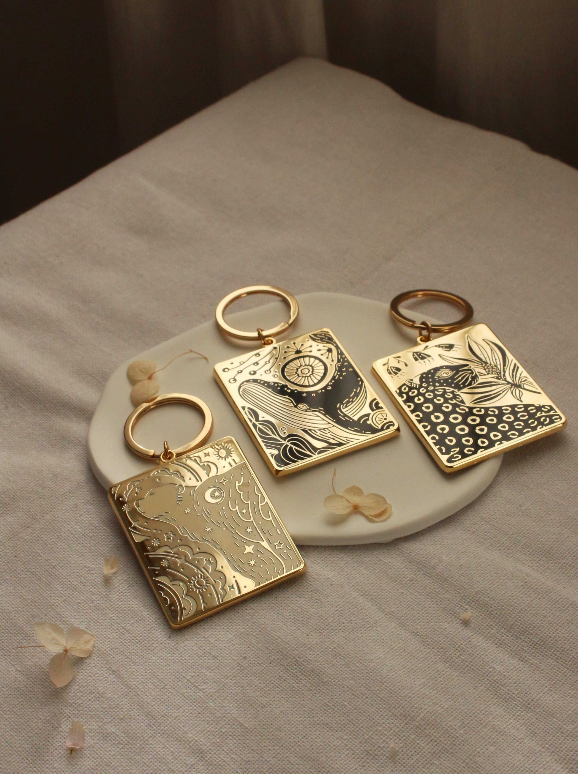 Set of three gold-plated enamel keychains with intricate designs displayed on a ceramic plate.