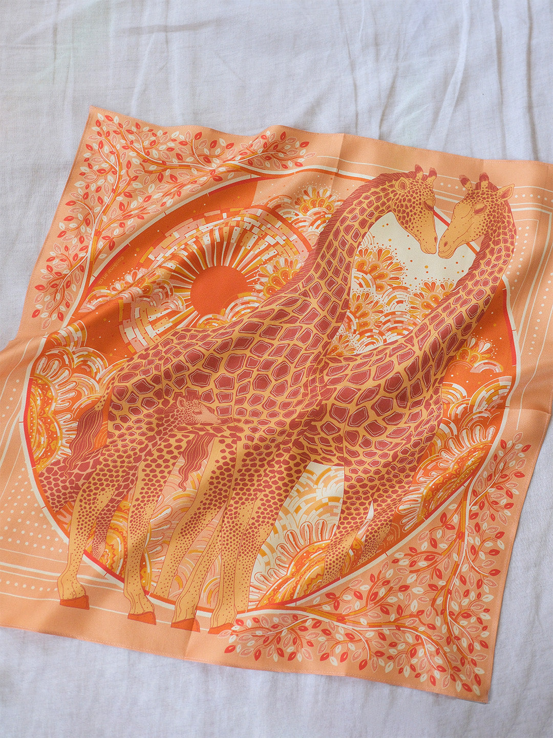 Scarf with a design of giraffes on an orange background, laid flat on a light gray surface.