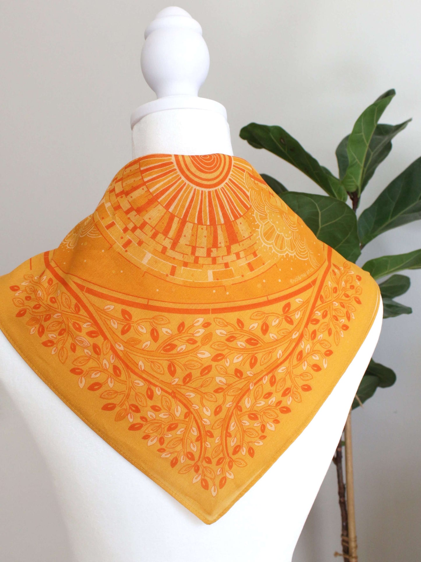 Golden yellow bandana scarf with intricate floral and geometric patterns, displayed on a mannequin against a green plant.