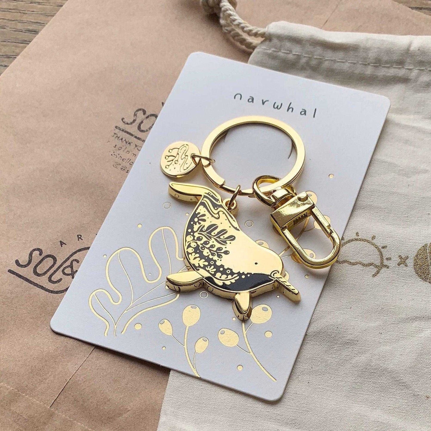Golden narwhal enamel keychain on a decorative display card featuring line art and habitat illustration.