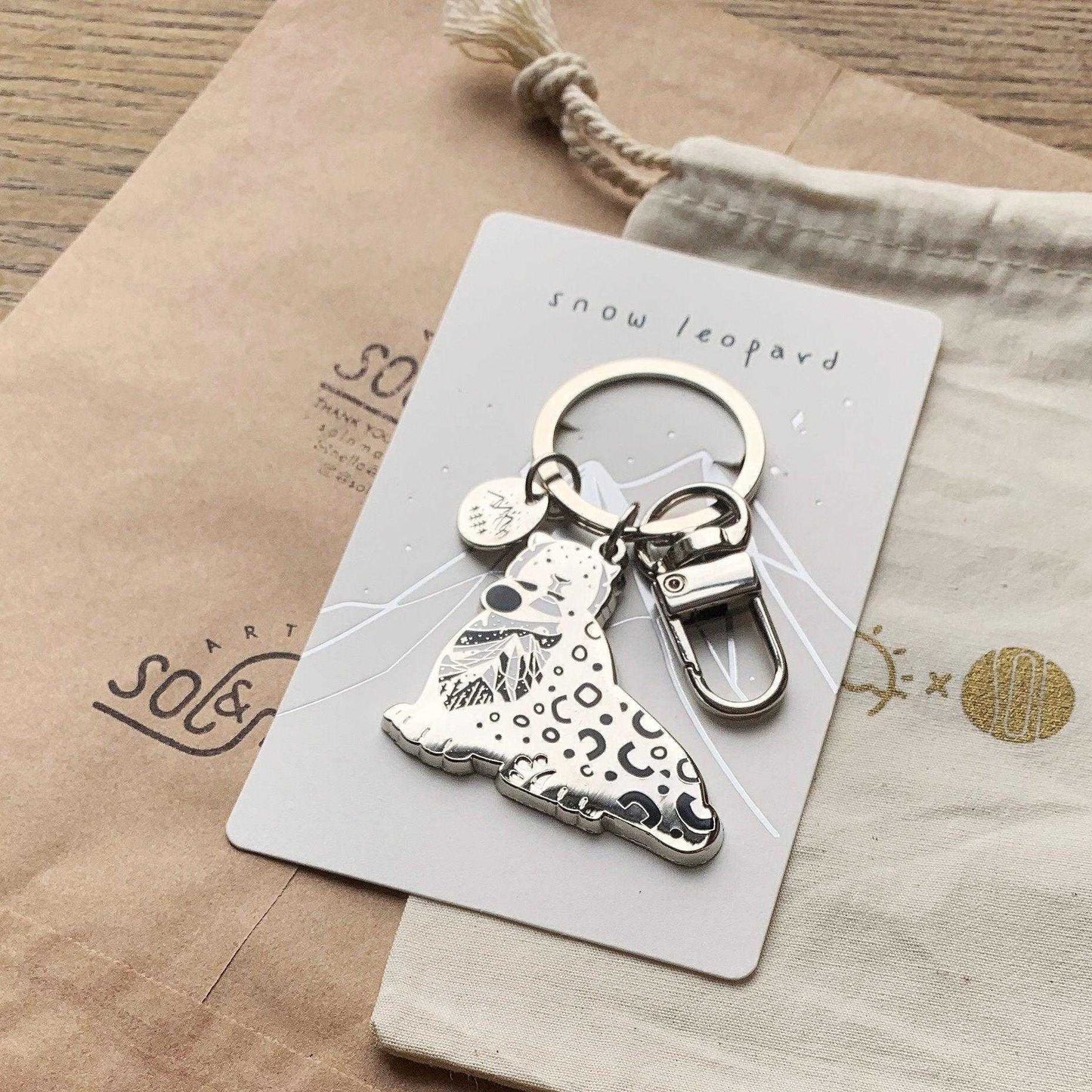 Snow Leopard enamel keychain on a display card, featuring its habitat and name, promoting animal conservation.