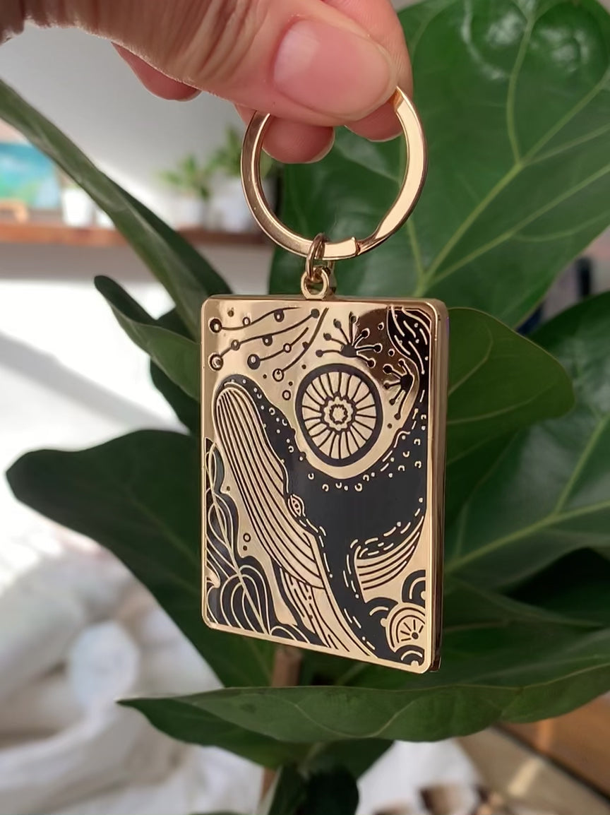 Humpback whale and ocean enamel keychain hanging on a plant, featuring a gold-plated design.