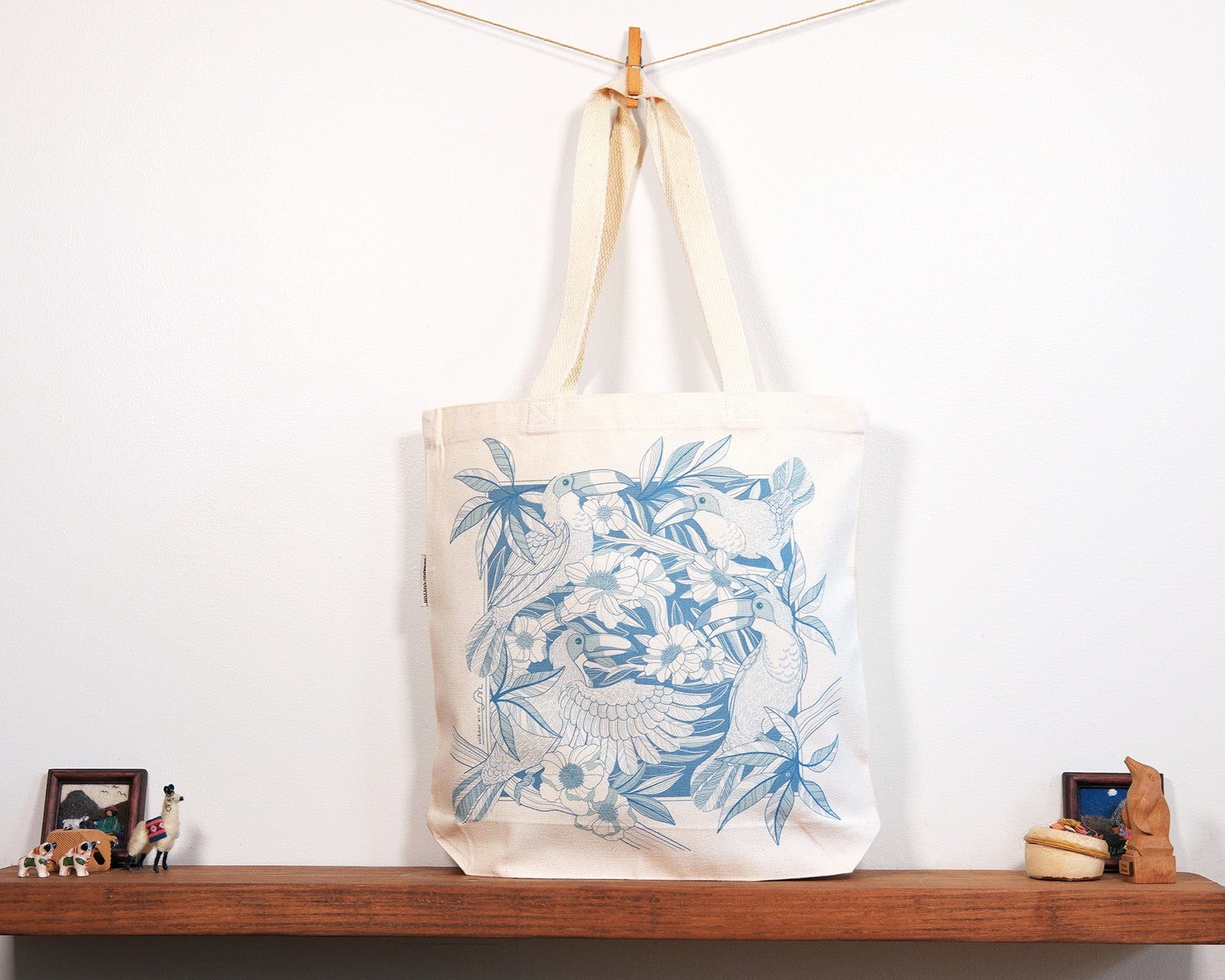 Tote bag with blue Toucan & floral design hanging on a wooden shelf against a white wall.