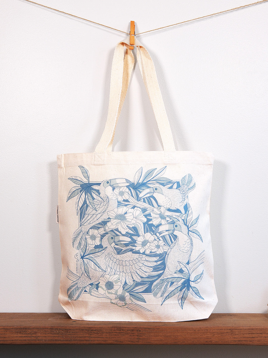 Tote bag with blue bird design hanging on a line against a white wall.