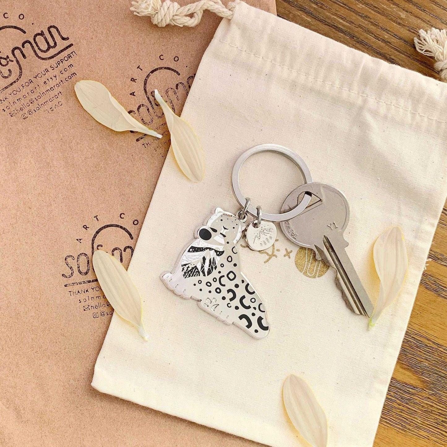 Snow Leopard enamel keychain design featuring habitat art, displayed with keys on a natural fabric pouch.