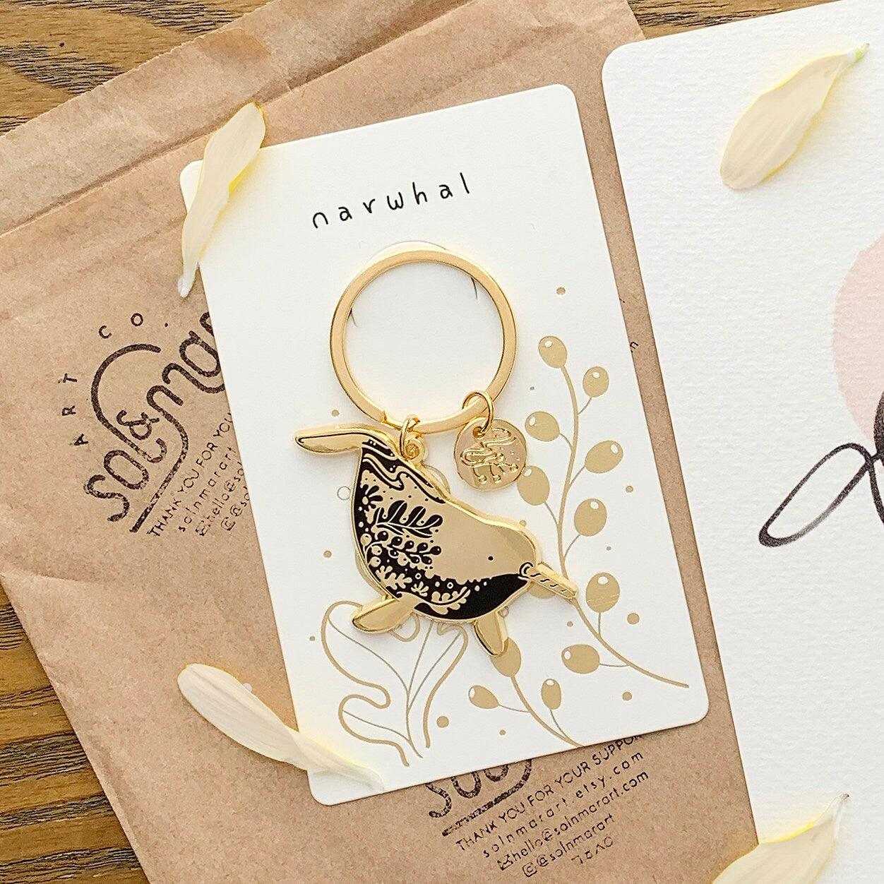 Narwhal enamel keychain on display card showcasing its habitat art, promoting endangered species awareness.