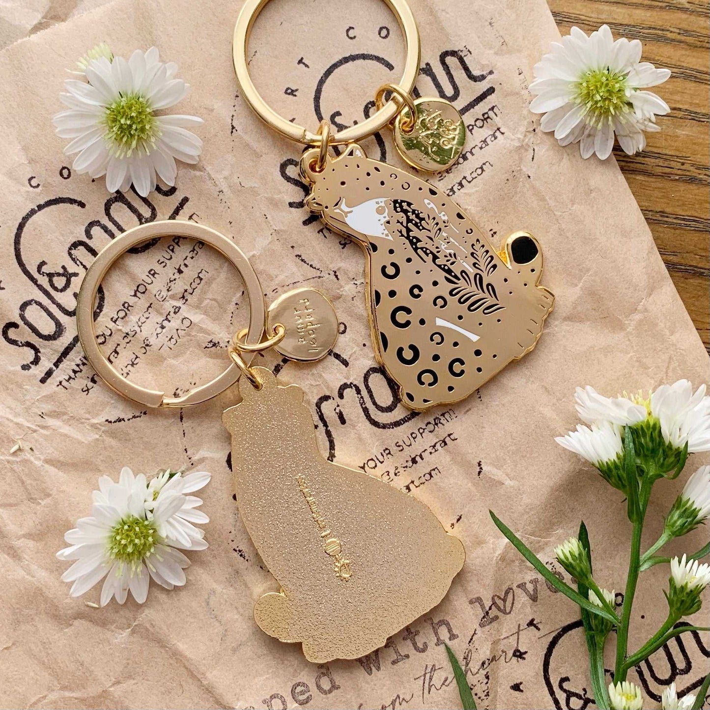 Amur Leopard enamel keychain with habitat illustration, adorned with delicate flowers and eco-friendly display card.
