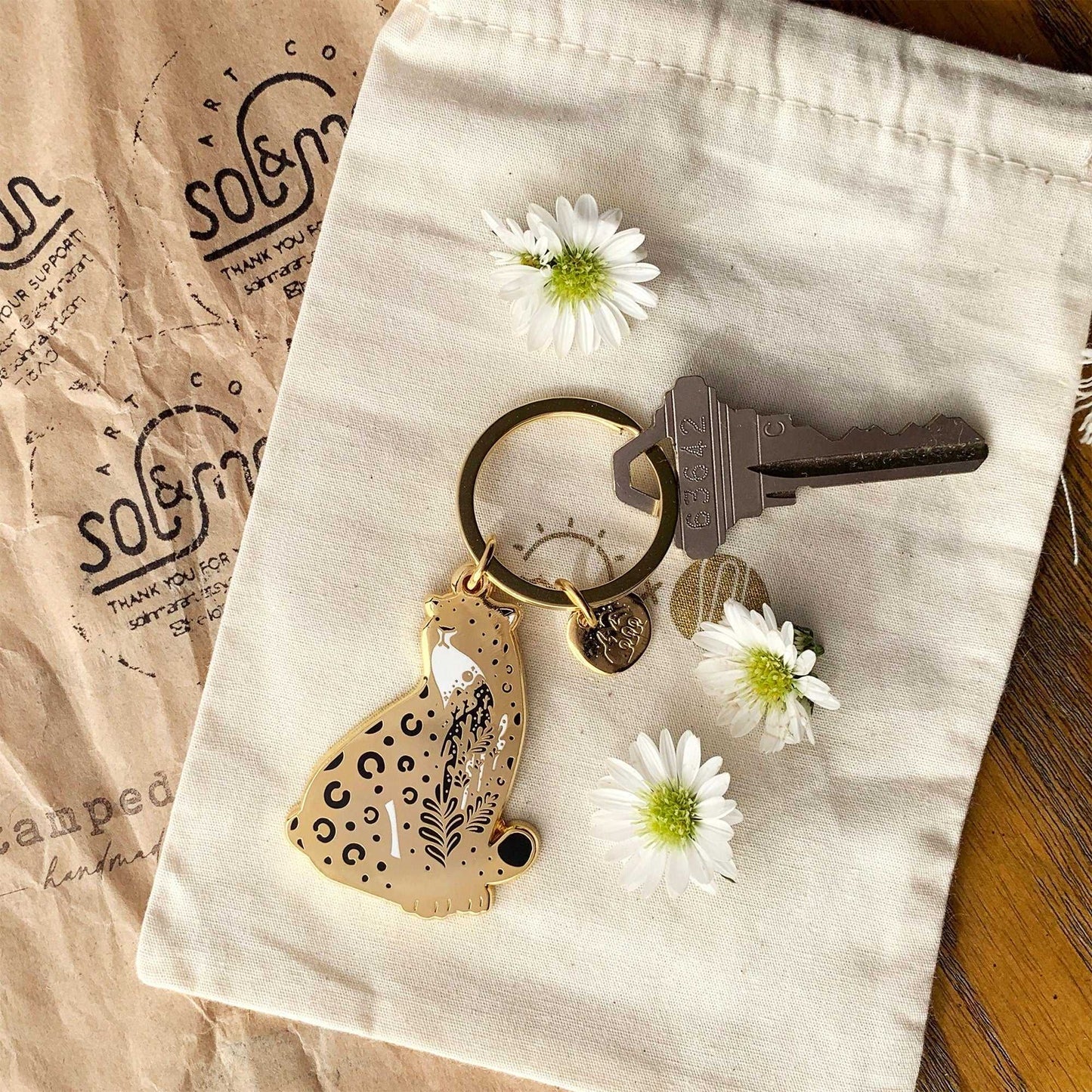 Amur Leopard enamel keychain on natural fabric with daisies and a key, promoting endangered species awareness.