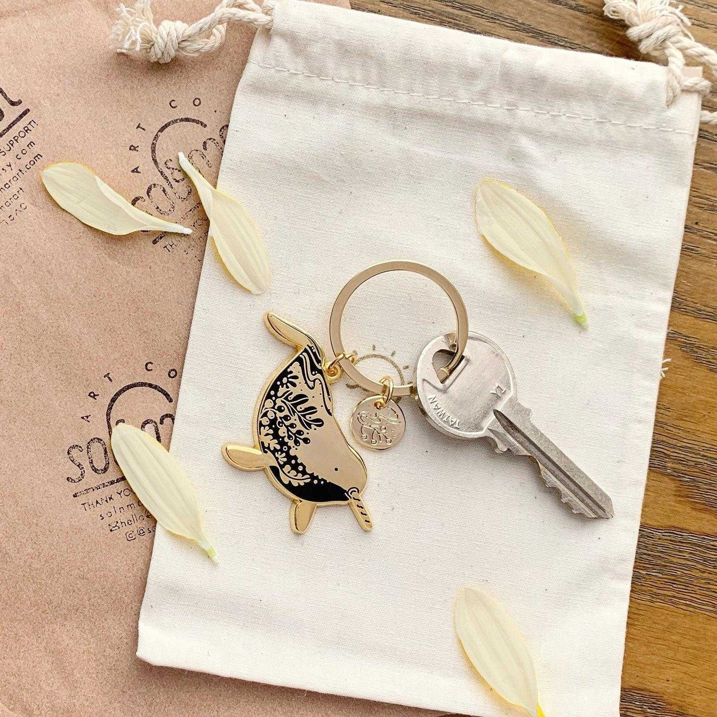 Narwhal enamel keychain in a drawstring bag, showcasing endangered species art and a circular charm, with a key.