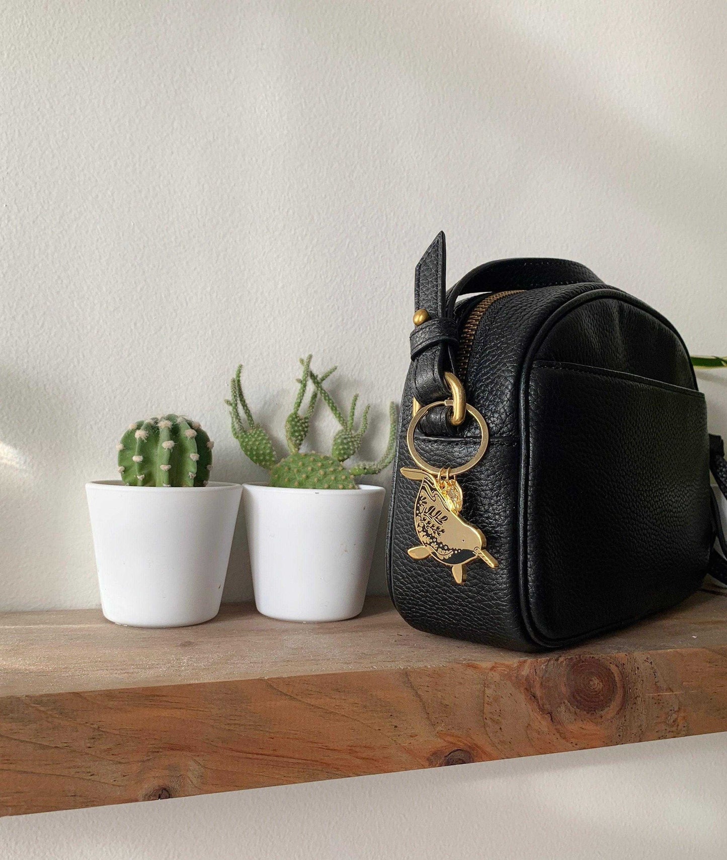 Black textured bag with narwhal enamel keychain on wooden shelf next to potted cacti.