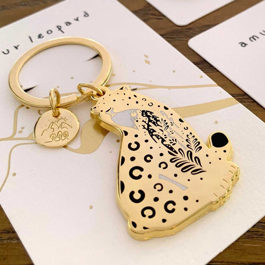 Amur leopard enamel keychain with habitat art and round charm on display card, promoting endangered species awareness.