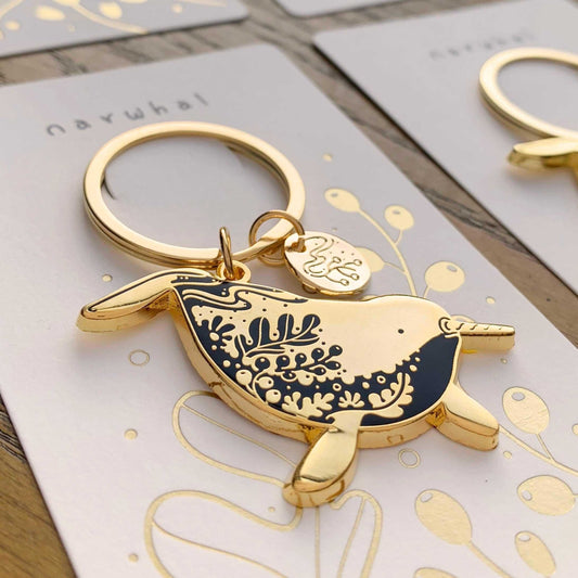 Gold narwhal enamel keychain featuring delicate habitat art, displayed on a decorative card, supporting endangered animals.