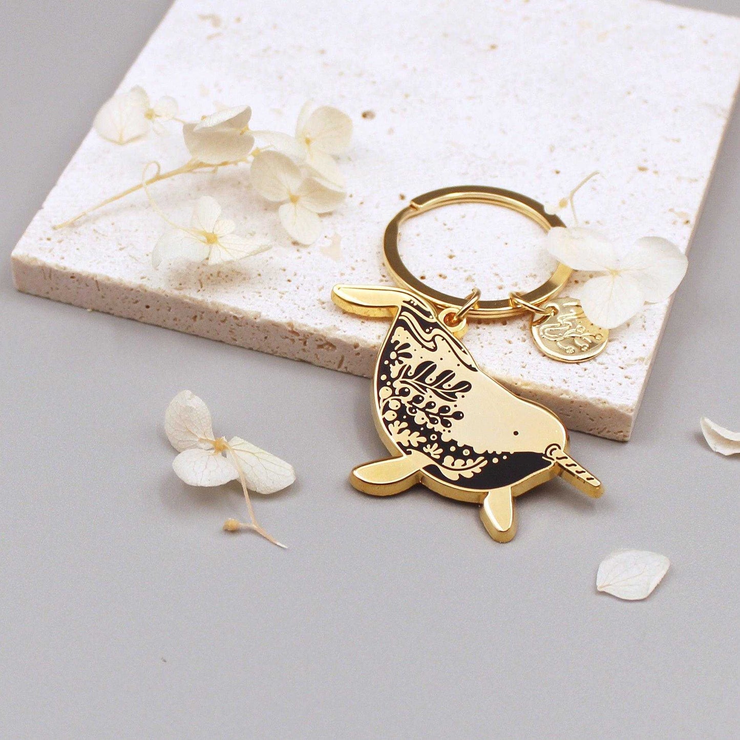 Gold narwhal enamel keychain with floral design, showcasing endangered species art on a stone background.