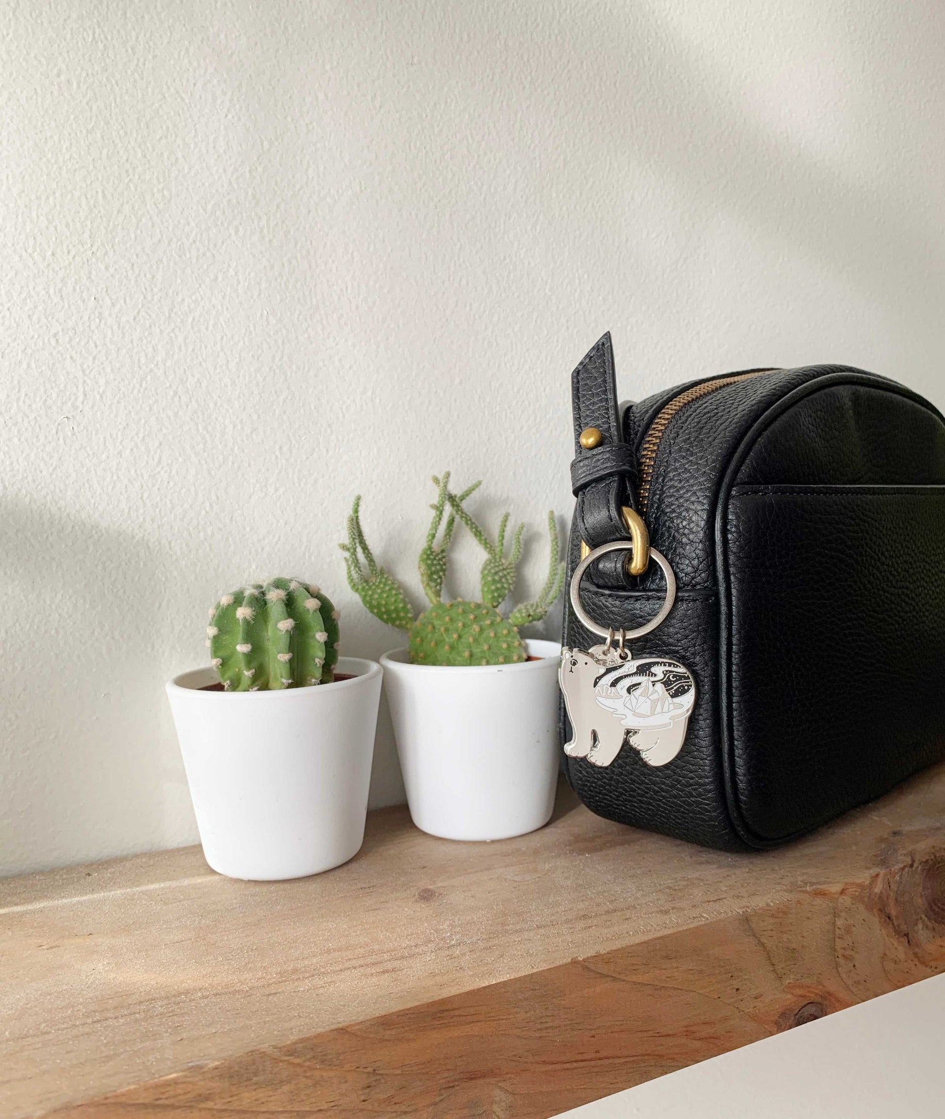 Black bag with Polar Bear keychain beside small potted cacti on wooden shelf.