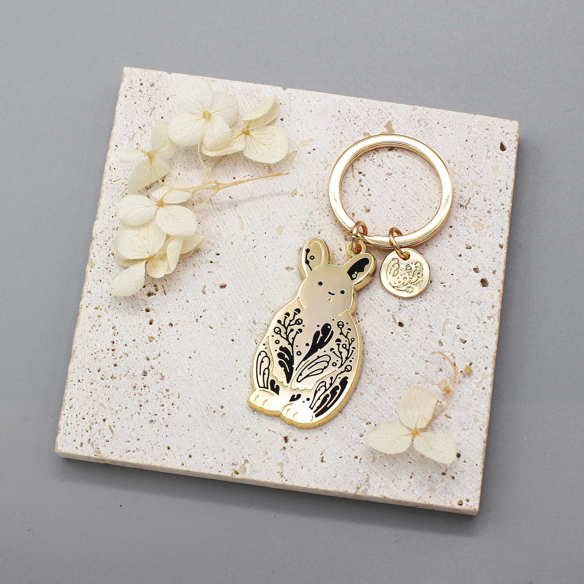 Pygmy rabbit keychain featuring floral design on gold charm, displayed on a textured background with dried flowers.