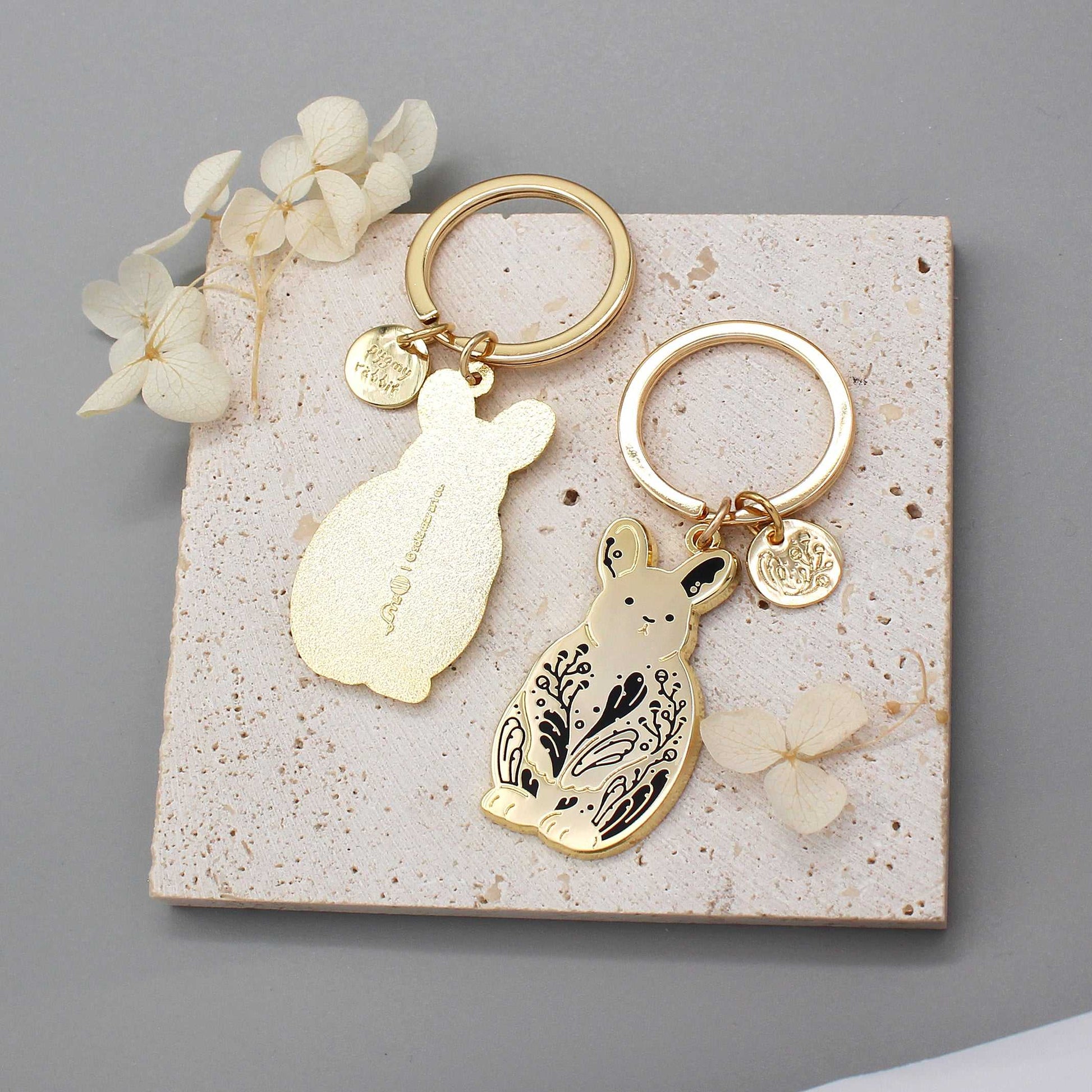 Gold bunny rabbit keychain set displayed on textured background with dried flowers.