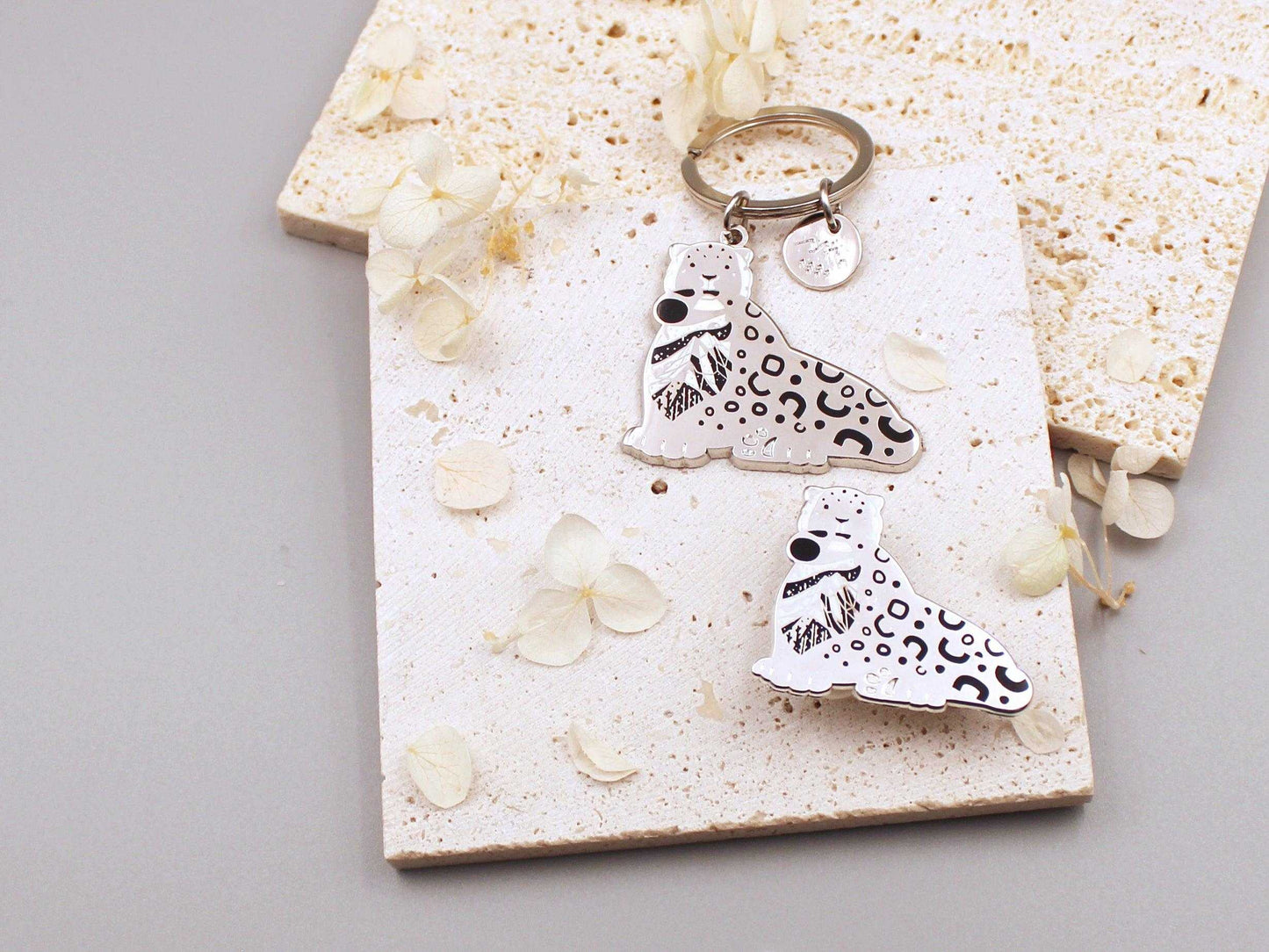 Snow Leopard enamel keychain on a textured display card, showcasing delicate habitat art and an organic tag.