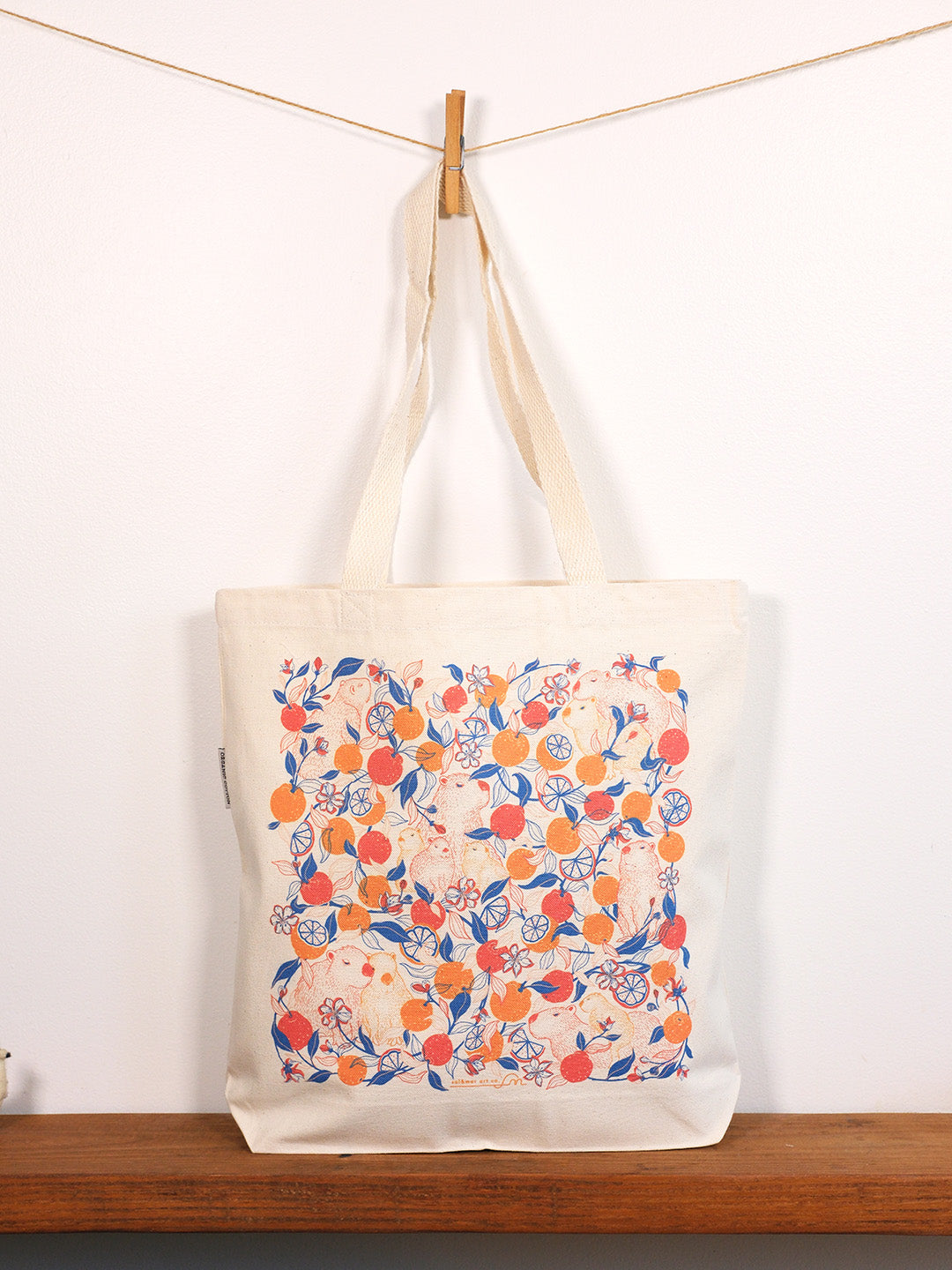 Tote bag with capybara & yuzu orange pattern hanging on a line against a white background