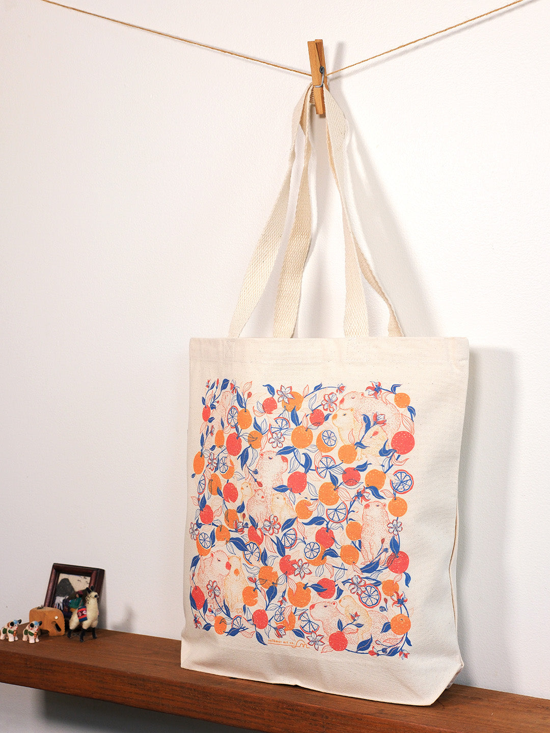 Tote bag with capybara & yuzu orange pattern hanging on a line against a white wall.