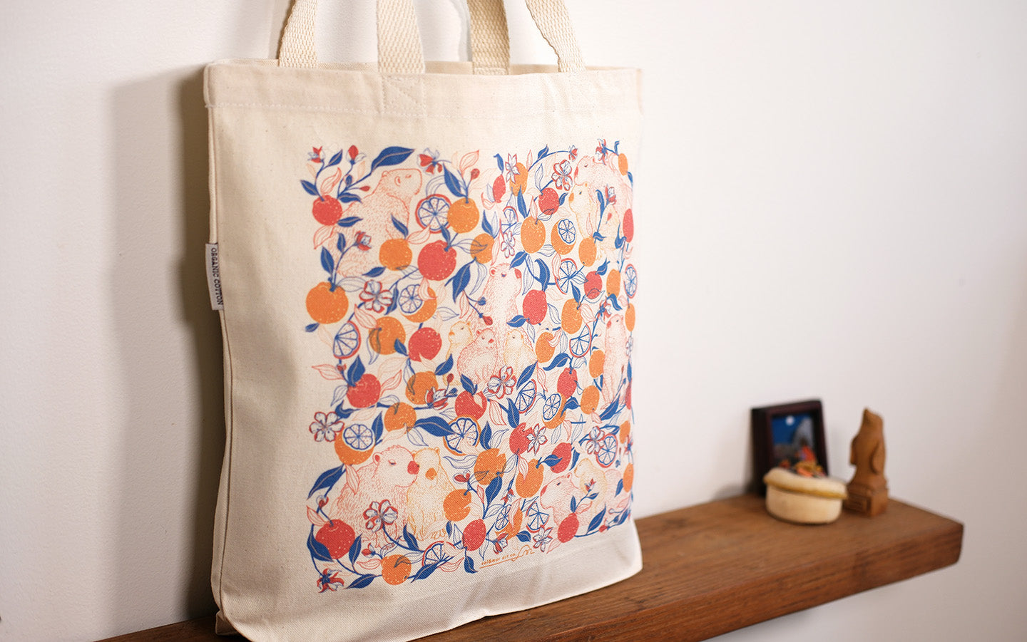 Beige tote bag with colorful capybara & yuzu orange pattern on a wooden shelf against a white wall