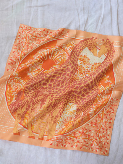 Scarf with a design of giraffes on an orange background, laid flat on a light gray surface.