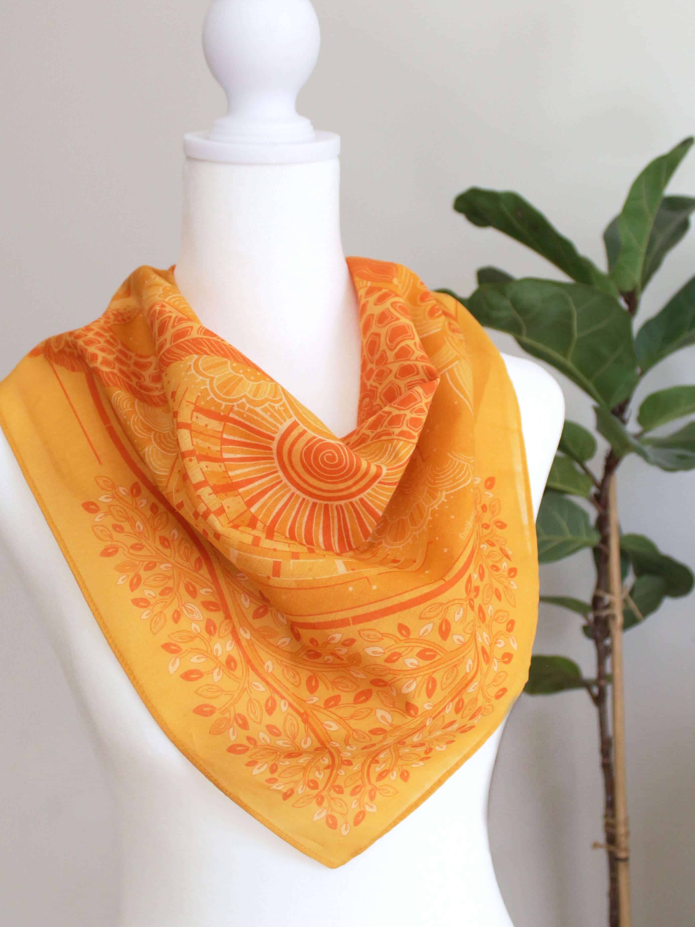 Golden yellow bandana scarf styled on a mannequin with vibrant sun and leaf patterns.