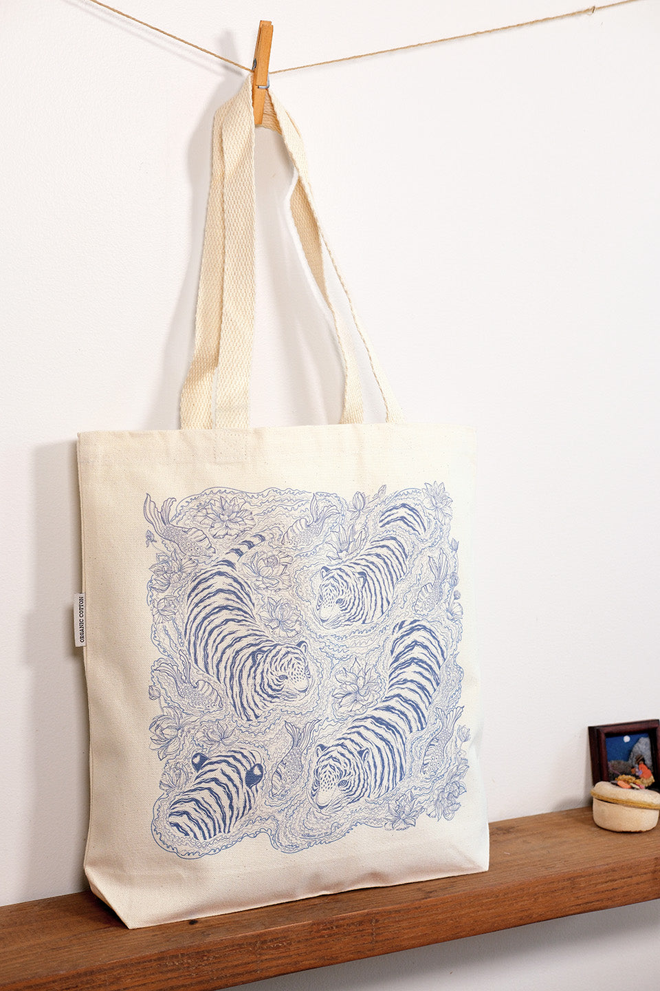 Tote bag with blue tiger design hanging on a wooden shelf against a white wall.
