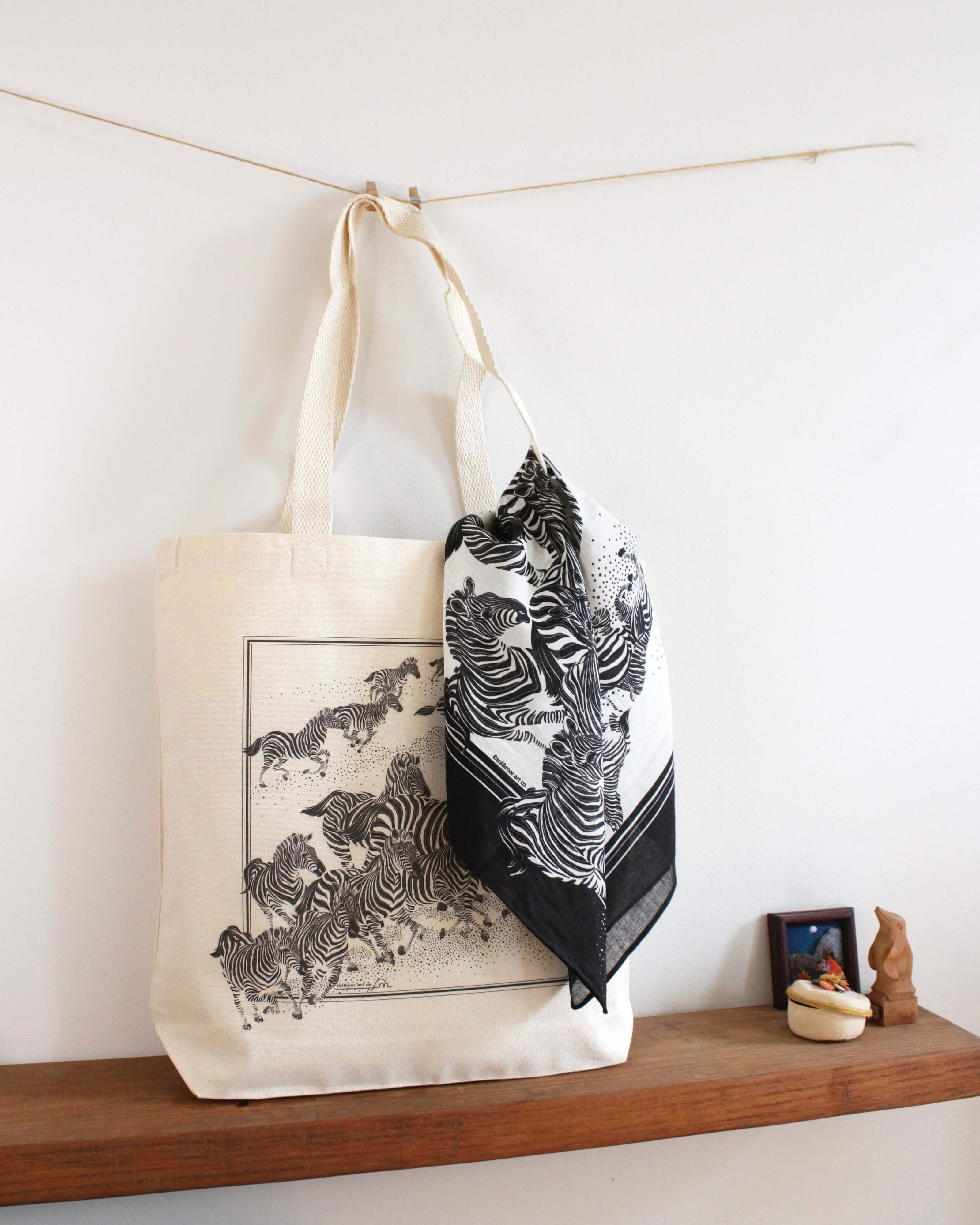Zebra "New Beginning" canvas tote bag with zebra illustration and black scarf displayed on a wooden shelf.